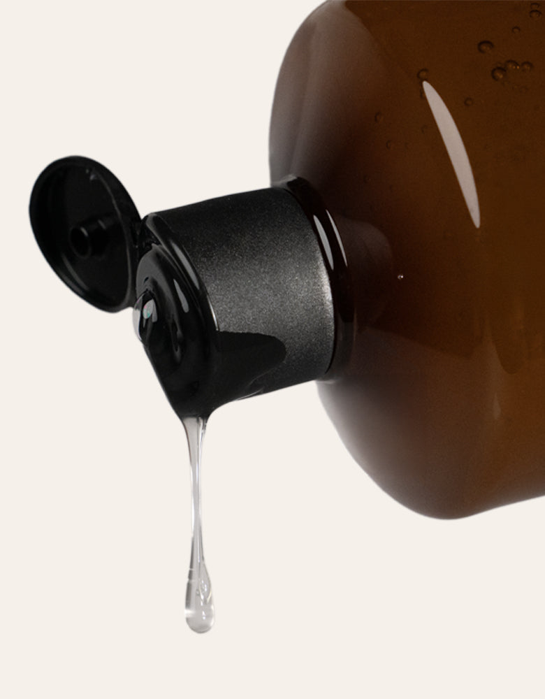 A drip of clear gel falling from the flip-top cap of an amber bottle of VIEVE Bath & Body Soak, on a cream background.