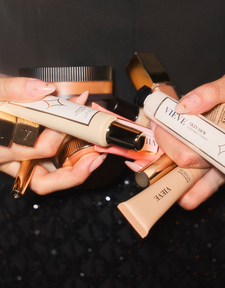 Hands holding a bundle of VIEVE best sellers against a dark, sparkly background, including Skin Dew, a Super Skin Nova jar, Skin Nova tubes and gold-capped makeup.