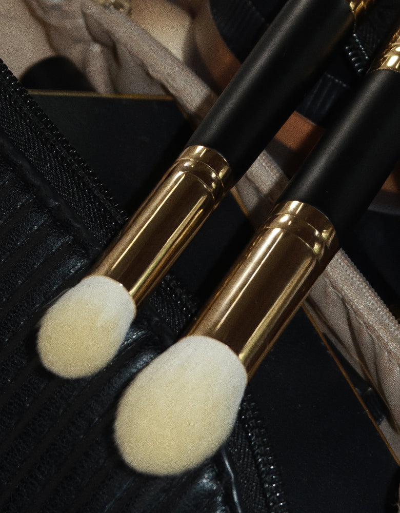 Two VIEVE makeup brushes with soft, domed bristles, black handles and gold ferrules, peeking out of a black makeup bag.