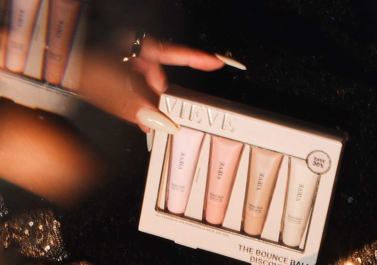 A hand holding The Bounce Balm Discovery Set by VIEVE: all four shades of the nourishing lip balm in mini size, in pink, coral, peach and cream tubes, against a dark, sparkly background.